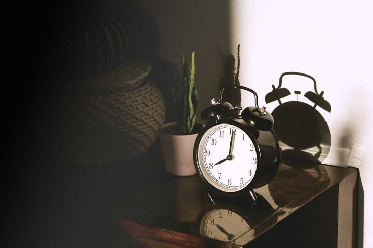 Black retro vintage alarm watch clock on a wooden bedside table with reflection in a polished surface in the rays of the setting sun with hard shadows